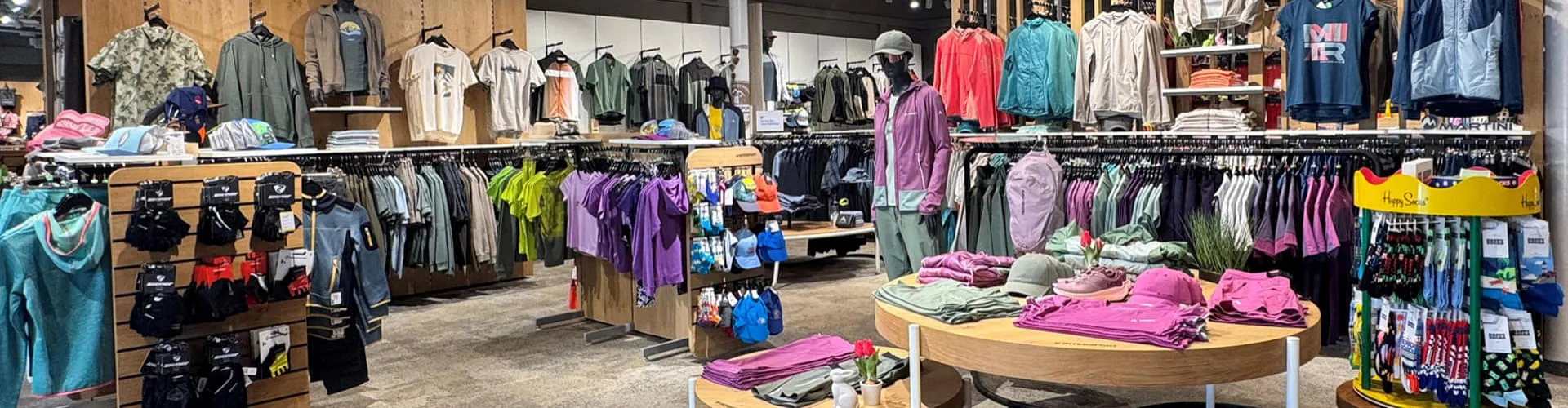 Das Bild zeigt den Innenbereich von INTERSPORT G&uuml;nther mit einer gro&szlig;en Auswahl an Sport- und Freizeitbekleidung, die auf Tischen, Kleiderst&auml;ndern und Regalen pr&auml;sentiert wird.